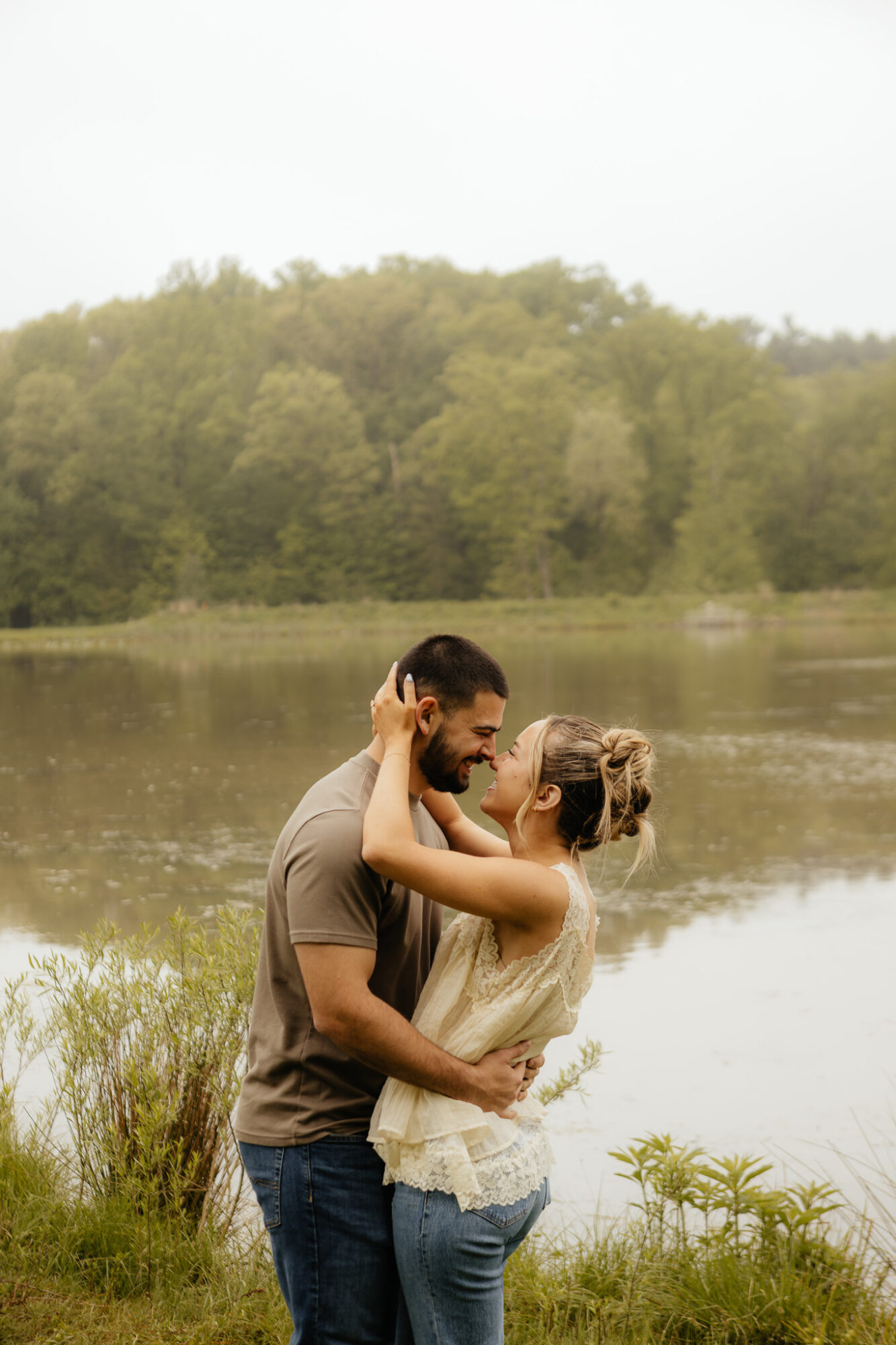 A couple stands by a lake, embracing and smiling at each other with trees and water in the background.