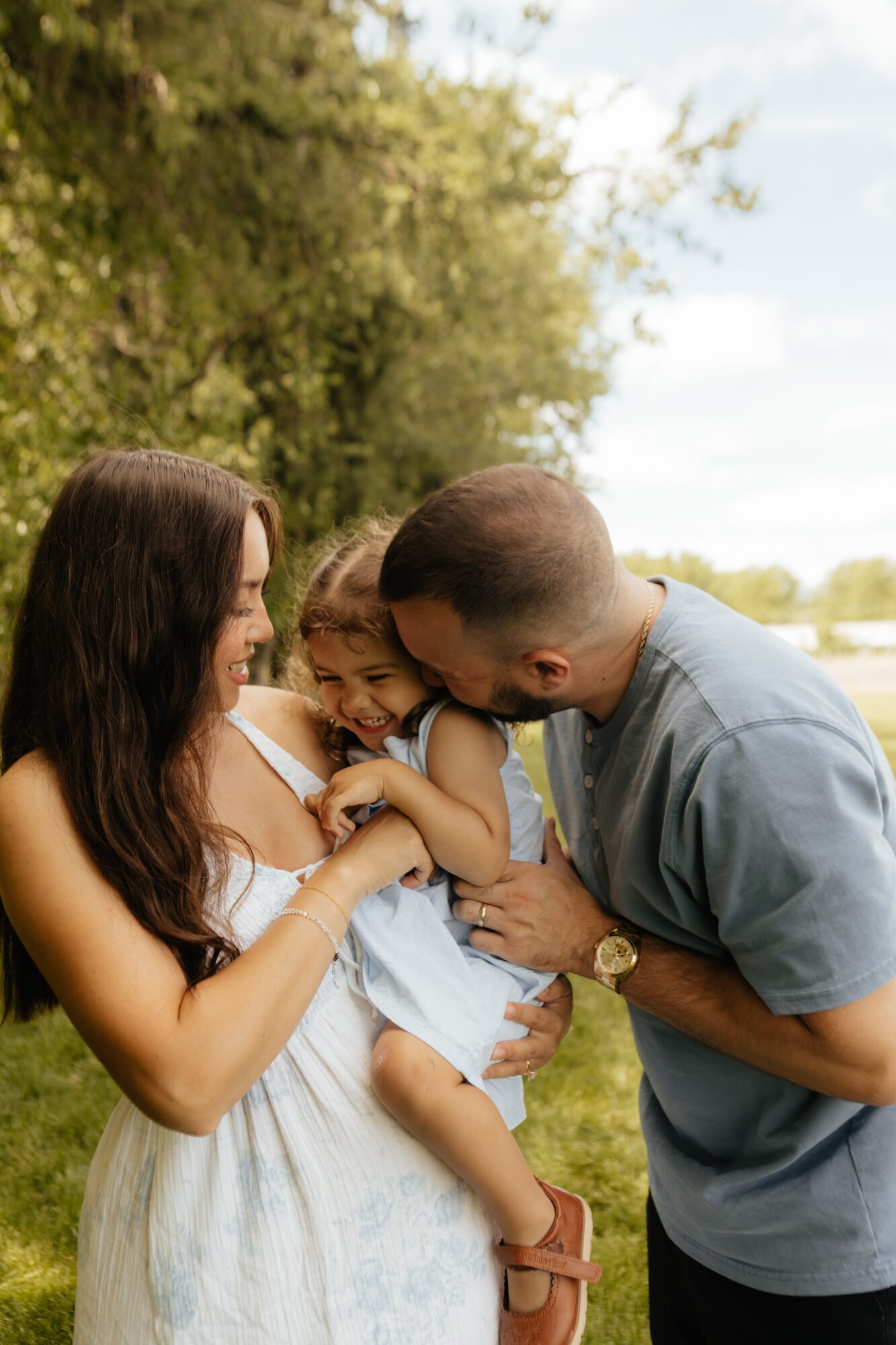 Family outdoors, woman holding child, man leaning in, trees and sky in background, sunny day.