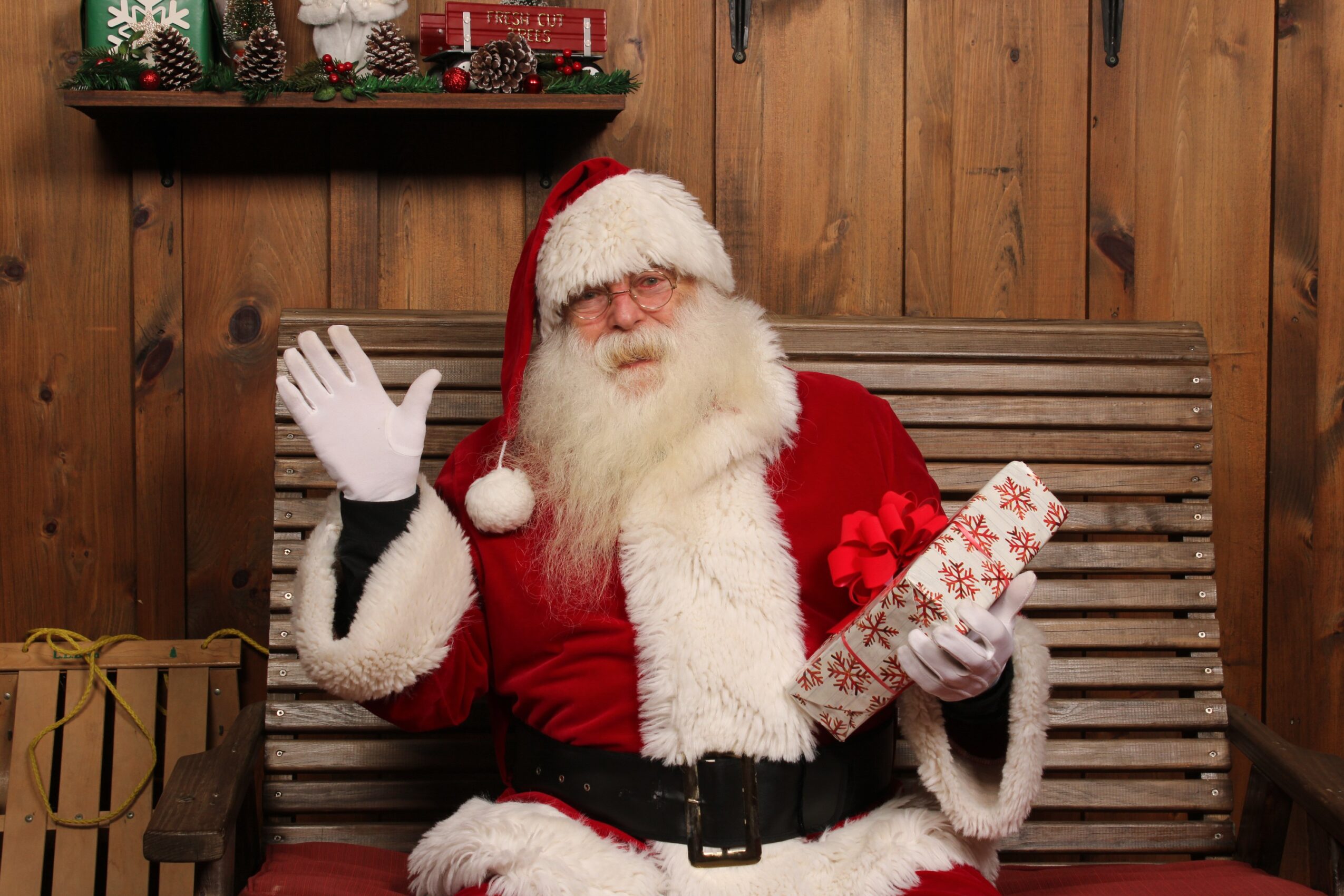 Santa Claus in a red suit with white trim, holding a wrapped gift, sitting on a wooden bench in a festive setting.