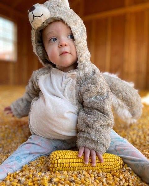 Child in animal hat and cozy jacket sitting on a carpet, holding a yellow corn cob, indoors with wooden walls.