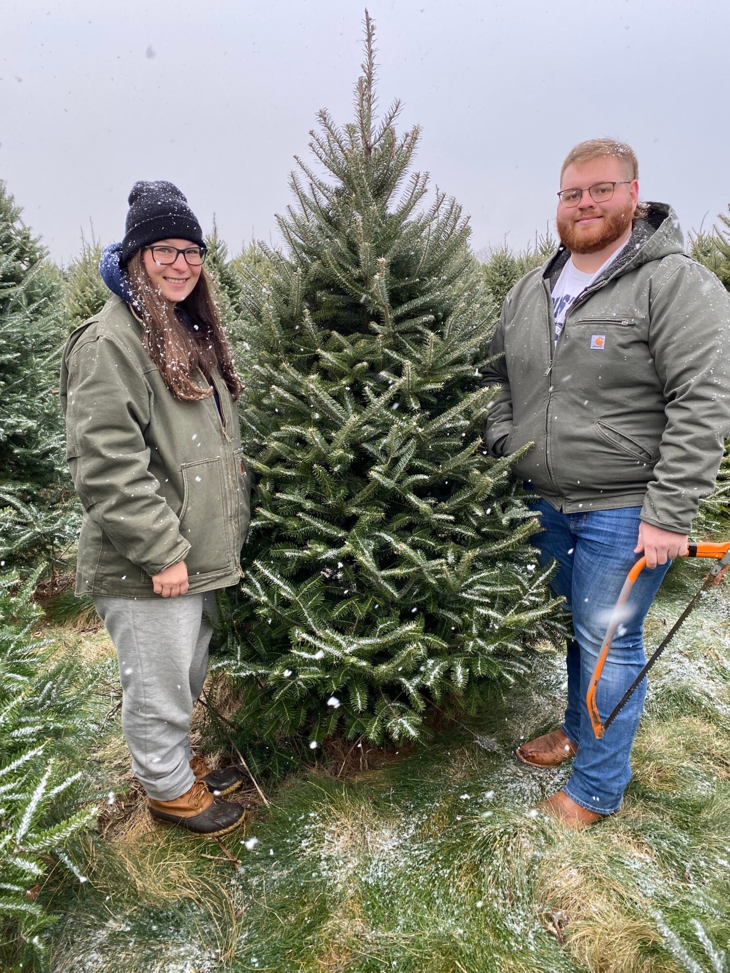 Two people stand outdoors next to a decorated Christmas tree, smiling, with grass and trees in the background.