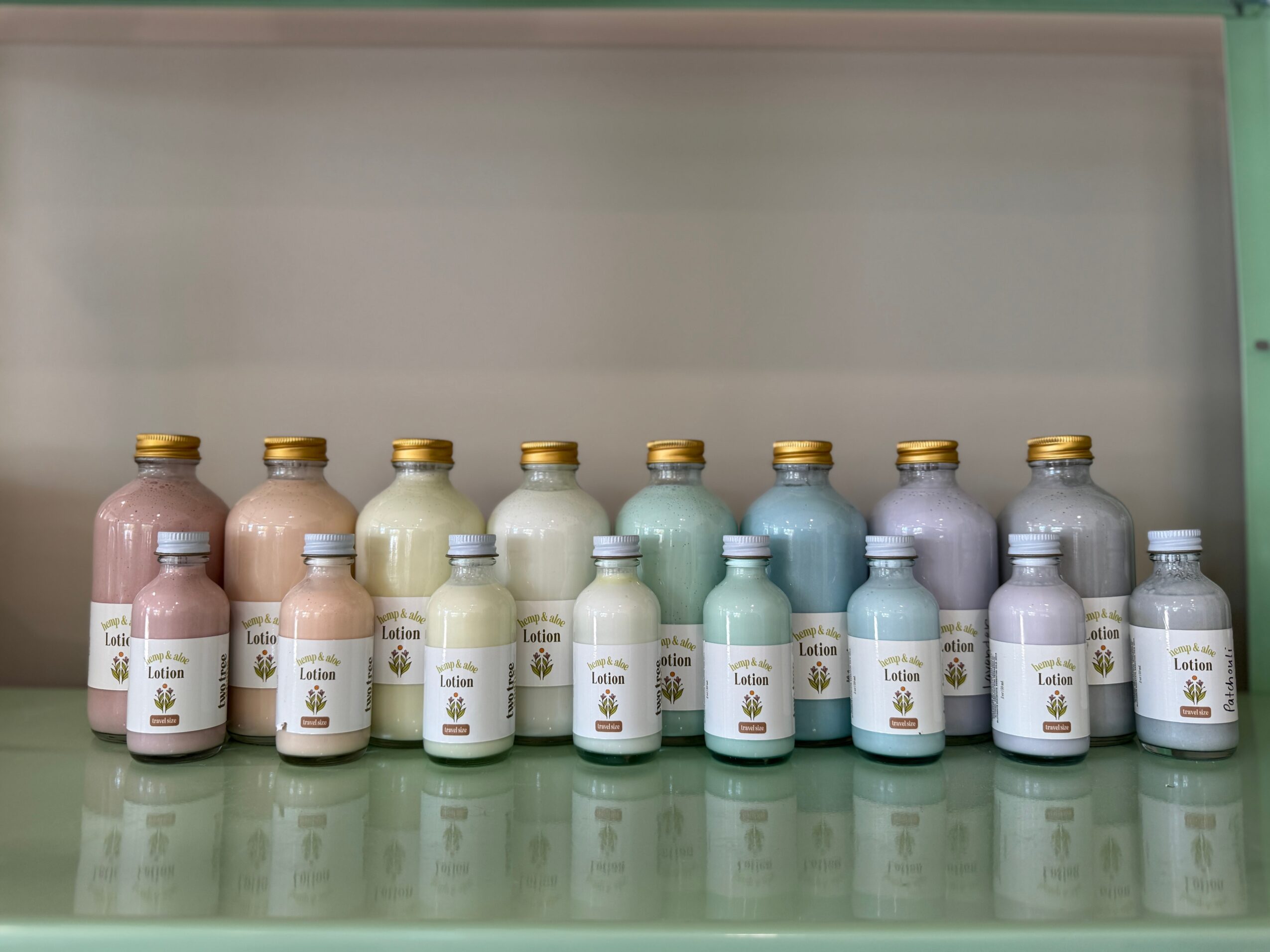 Row of twelve bottles with pastel-colored caps and labels on a reflective surface, against a plain background.