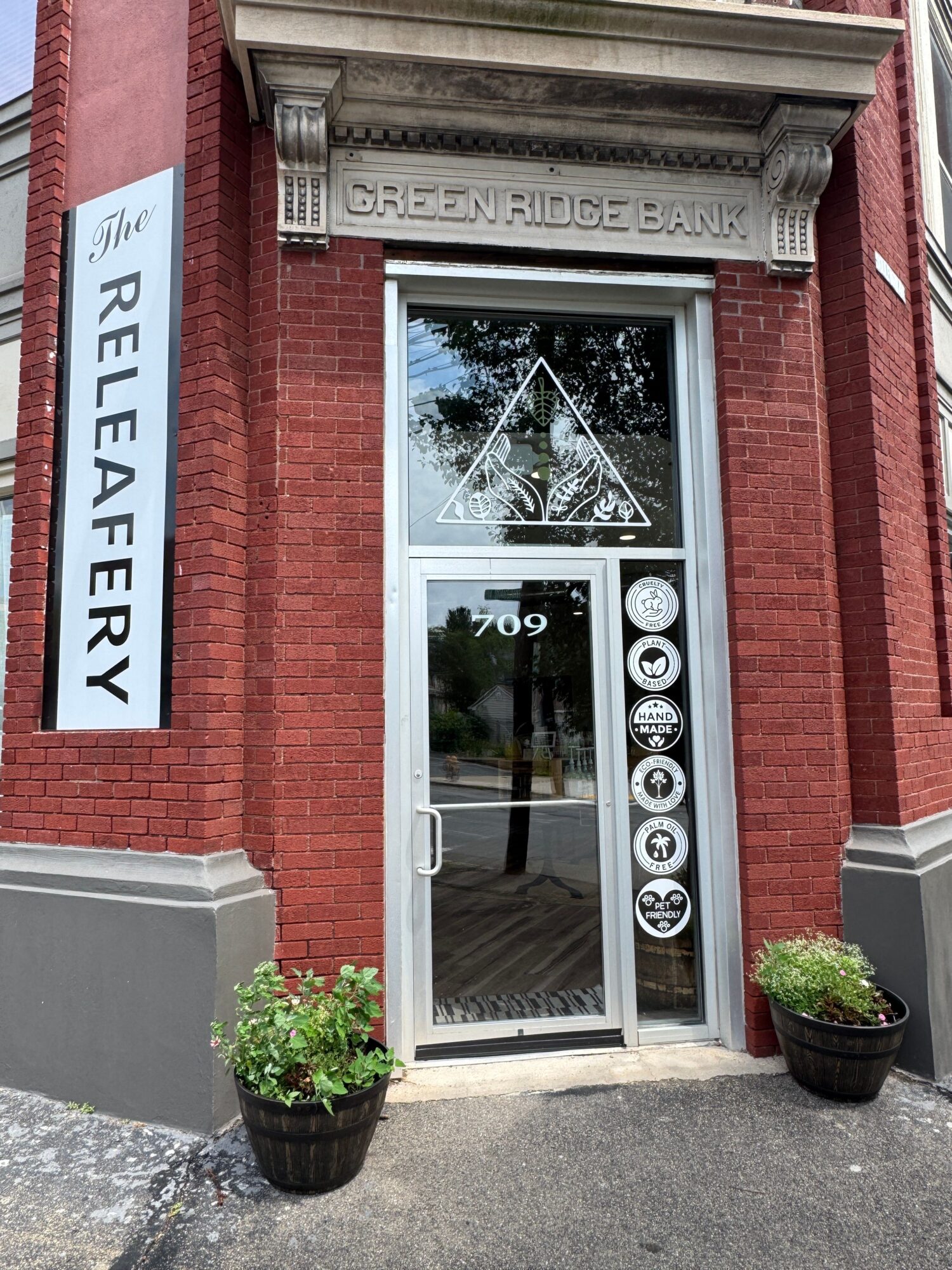 Red brick building with glass door and window, sign reads 'RELEAFERY', potted plants outside, reflection of trees in window.