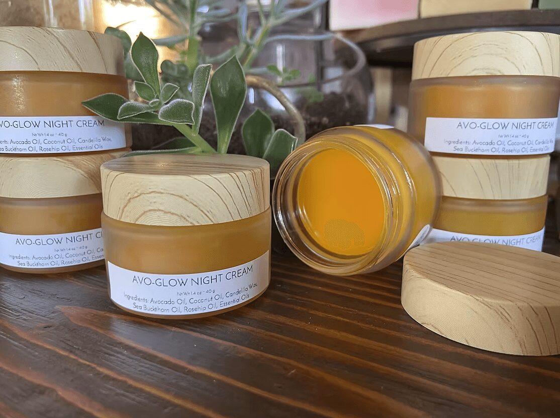 Several jars of night cream with wooden lids on a wooden surface, some jars are open, with a plant in the background.