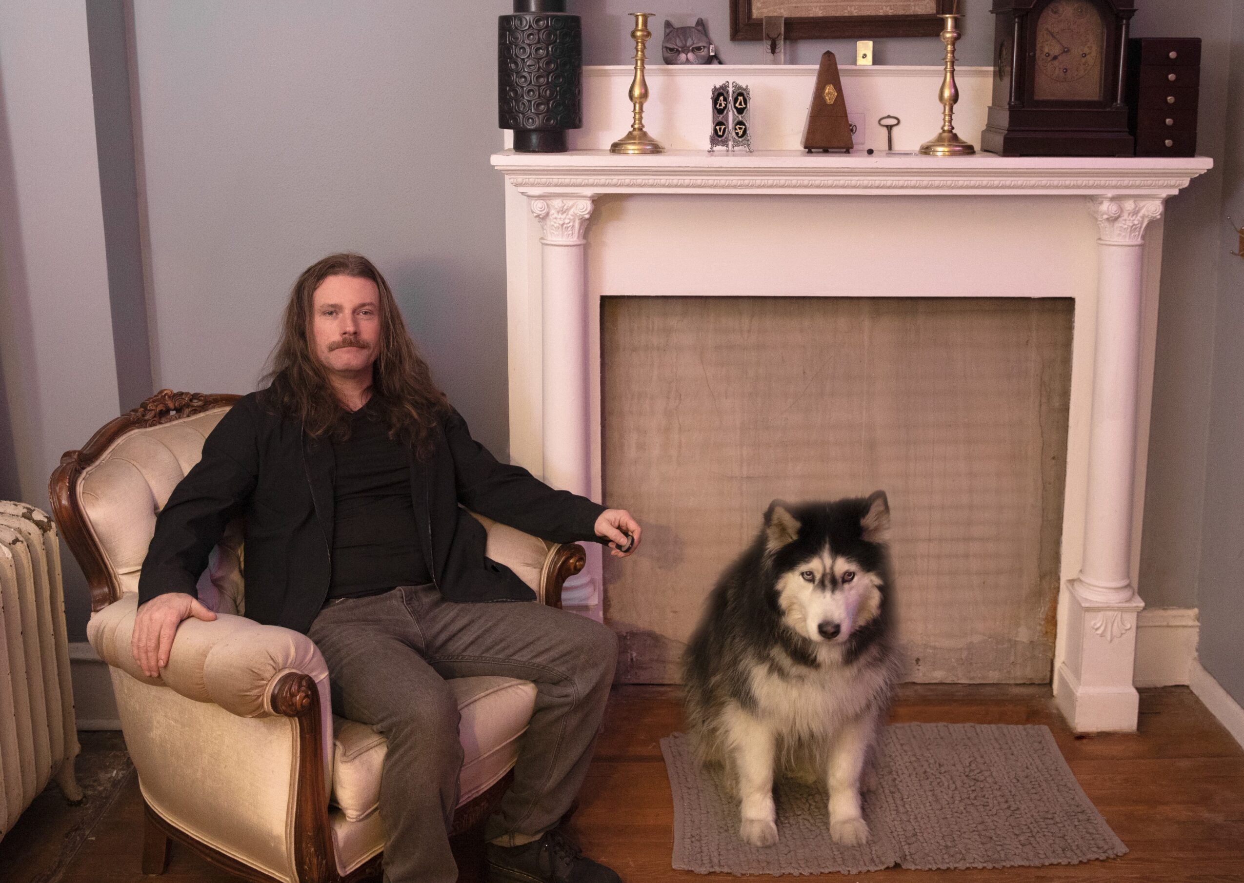 Man with long hair and beard sitting on a vintage armchair next to a large fluffy dog in front of a fireplace.