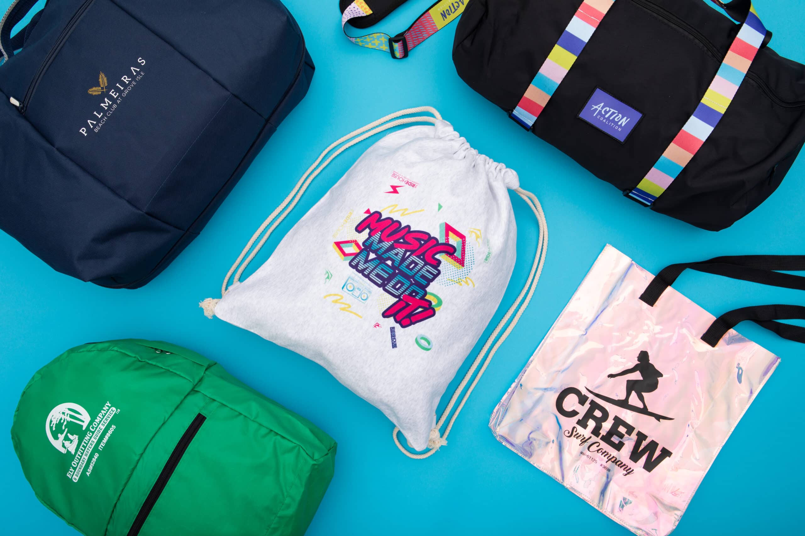 Three bags and a drawstring bag on a blue surface, including a navy backpack, a black bag with colorful straps, and a pink bag with a running figure, and a green bag.
