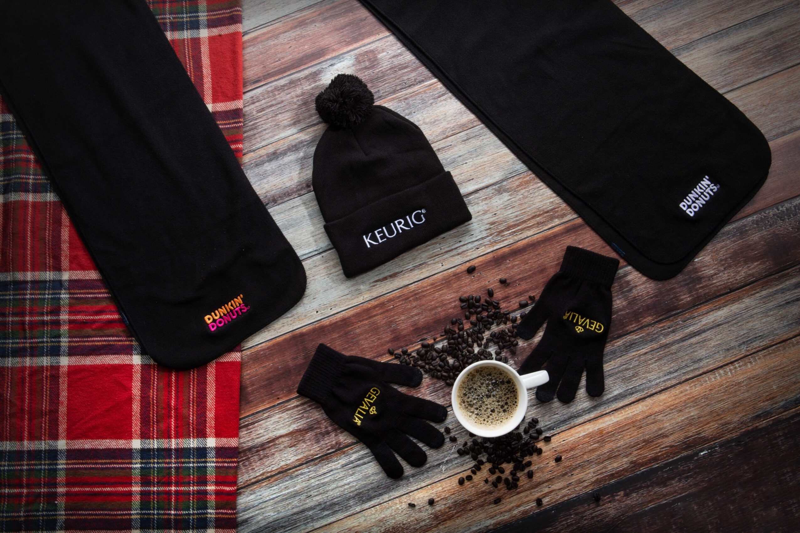 Keurig coffee cup with coffee beans, gloves, a beanie, and two black cases on a wooden surface, with a red plaid cloth partially visible.
