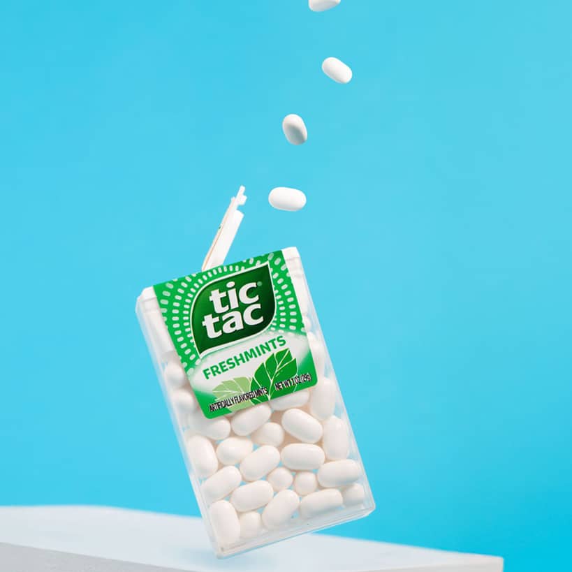 A pack of Tic Tac mints with some mints falling into the air against a blue background.