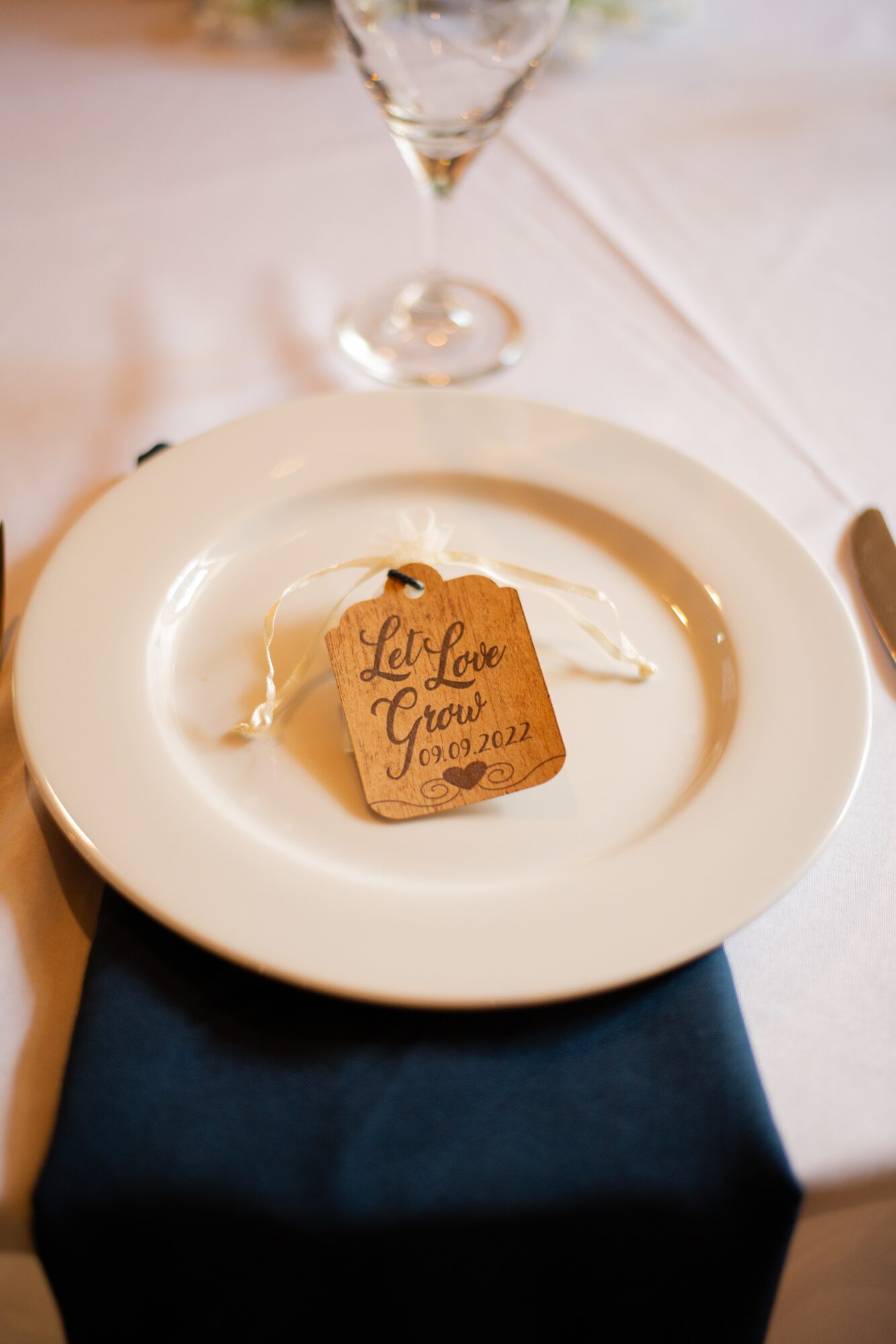 A white plate with a small wooden plaque on it, placed on a table with a white tablecloth and a glass in the background.