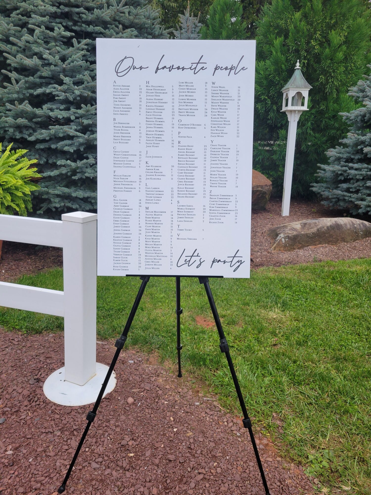 White signboard with text, mounted on black tripod, outdoors on a gravel path, surrounded by green grass and trees.