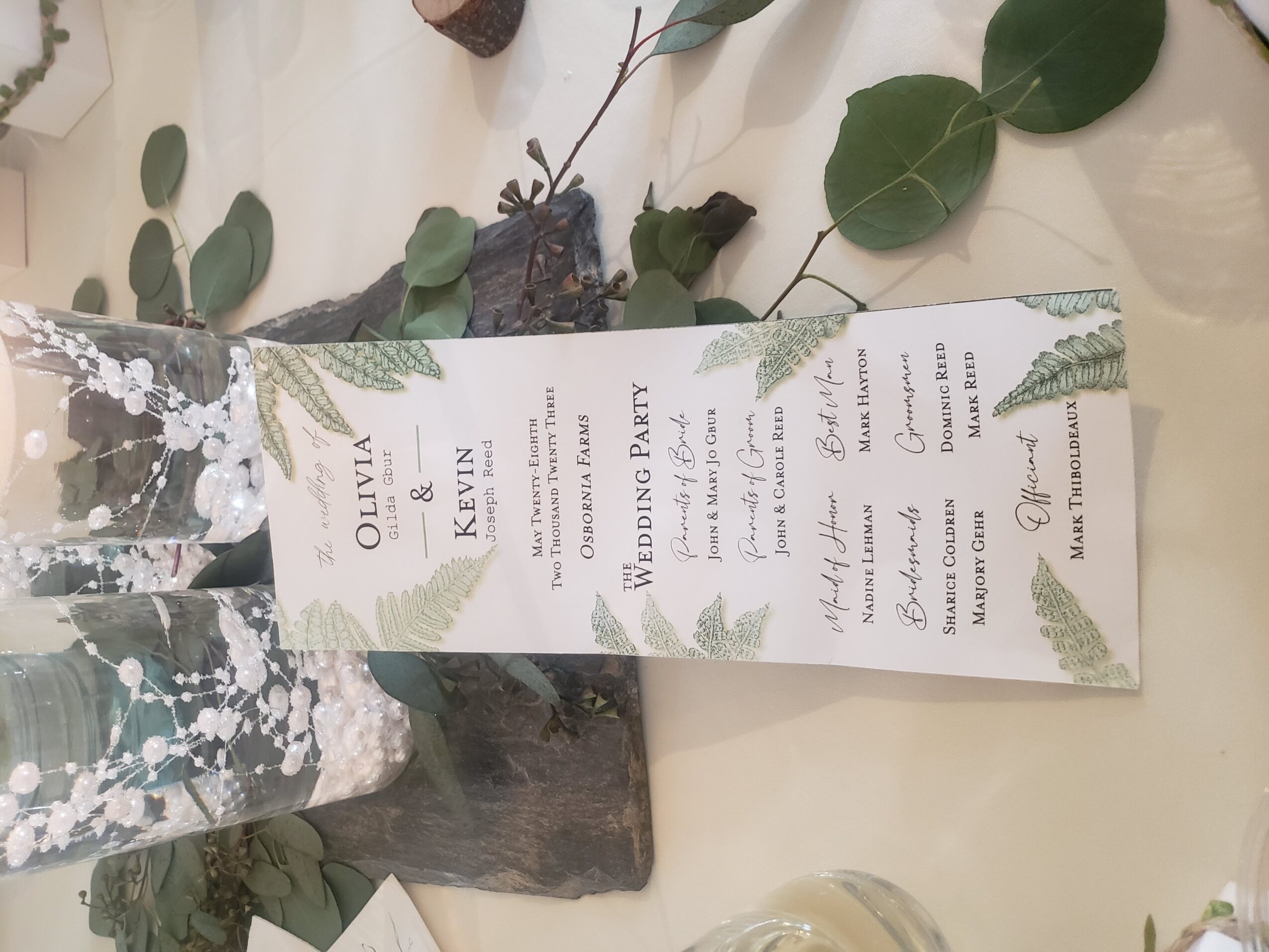 Wedding program with green fern decorations, listing names Olivia and Kevin, and event details, on a table with leaves and a candle.