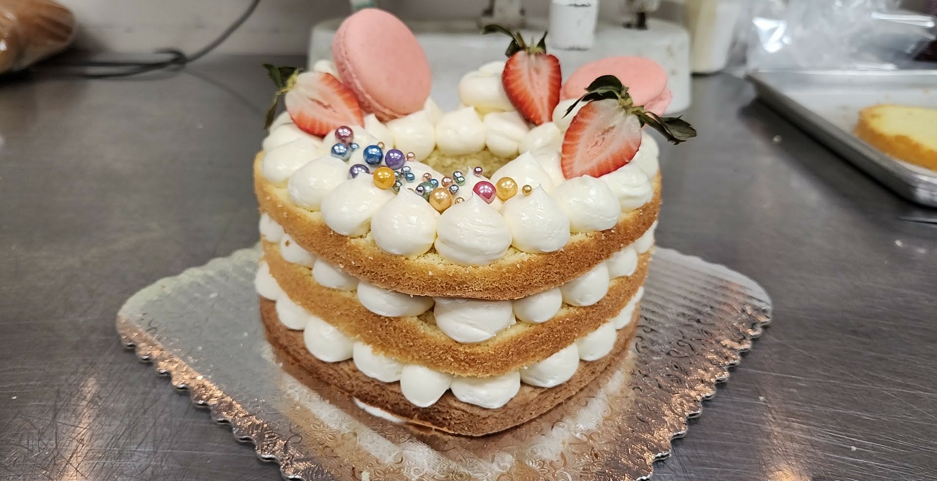 Three-layer heart-shaped cake with white frosting, strawberries, macarons, and colorful sprinkles, on a silver surface.