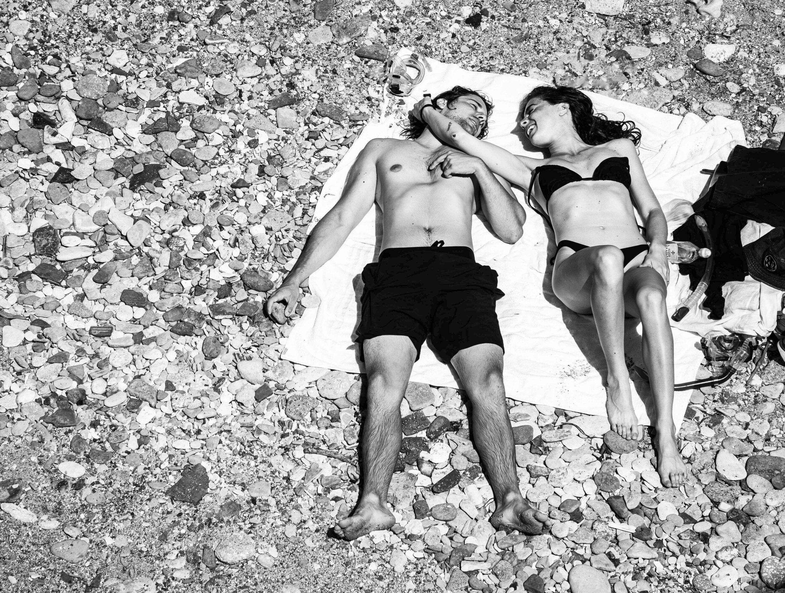 Two people lying on a towel on rocky ground, one appears to be a man and the other a woman, holding hands.