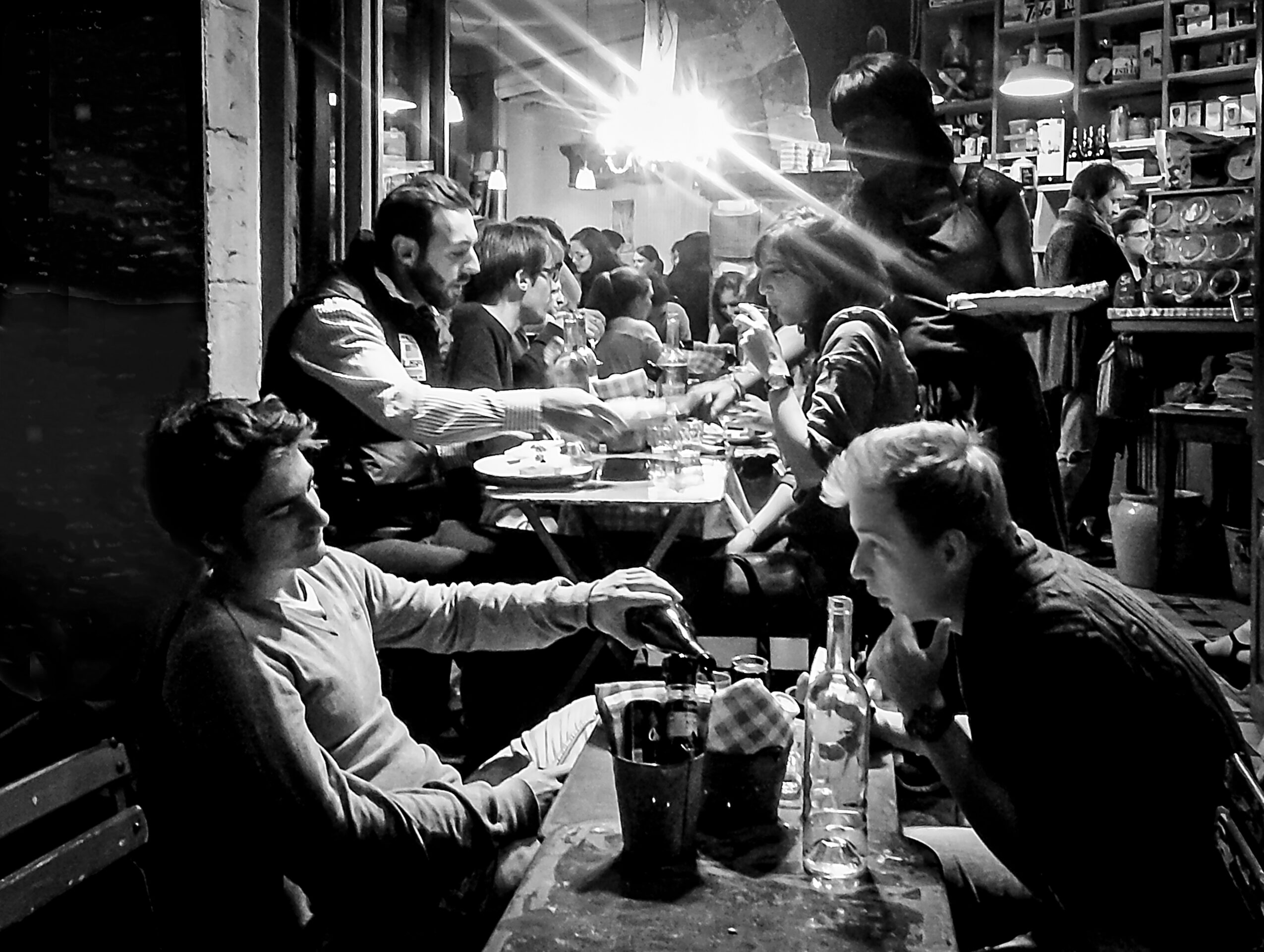 People dining at tables in a busy restaurant with shelves and a server in the background.