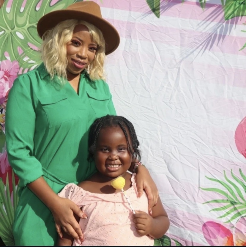 Woman and girl smiling, standing in front of a tropical-themed backdrop with leaves and pink flowers.