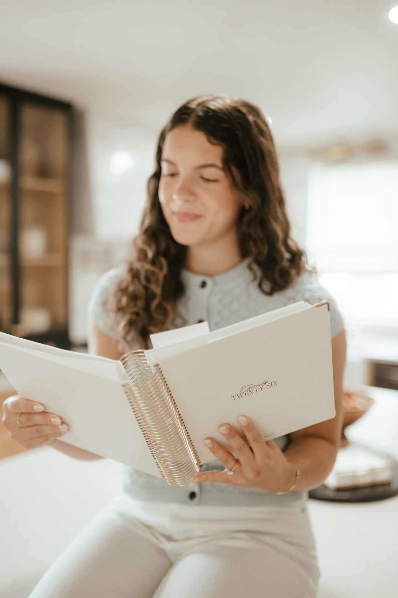 Young woman with long curly hair reading a notebook indoors, smiling gently, wearing a light-colored top and white pants.