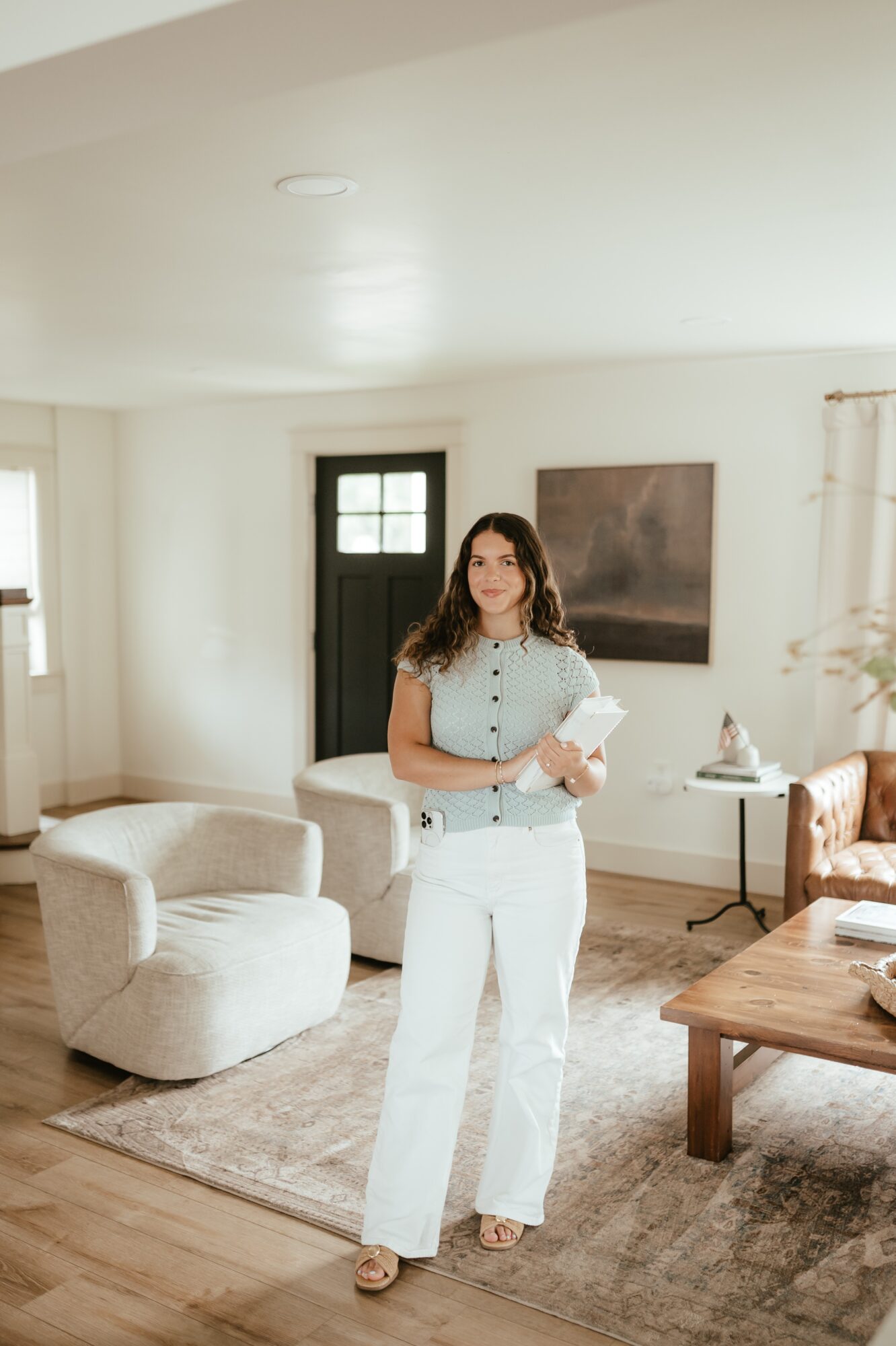 Woman standing in living room holding a book, with sofa, table, and window in background.
