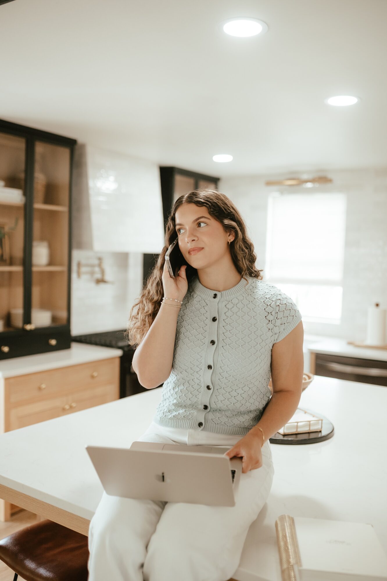 Woman with curly hair talking on phone in kitchen, holding laptop, bright room with large window, light-colored walls.