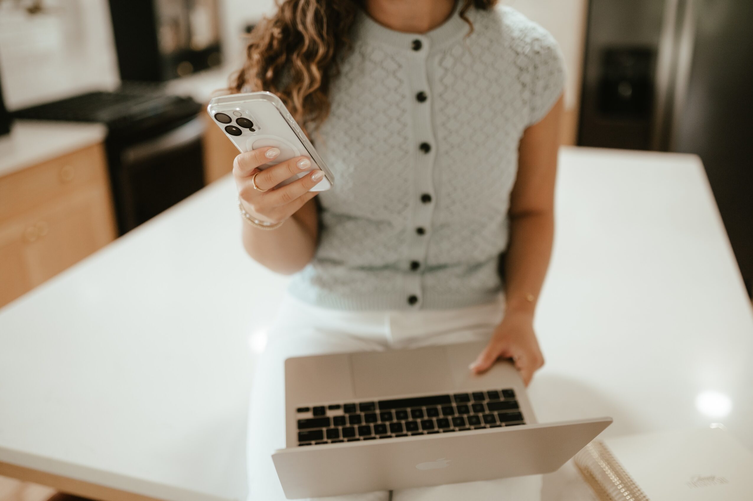Woman holding a smartphone in a kitchen, with a laptop on a white table in front of her.