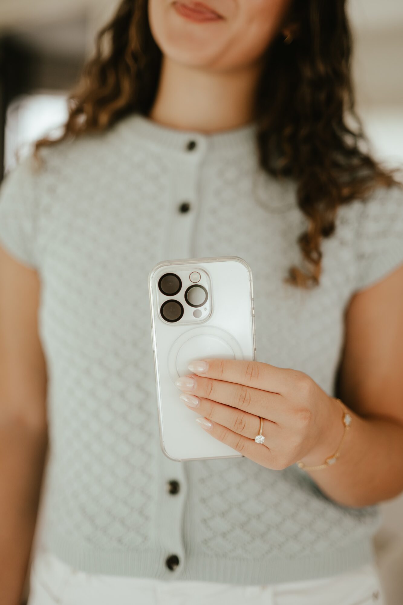 Woman taking a selfie with a smartphone, wearing a light-colored, textured blouse, with curly hair and a ring on her finger.