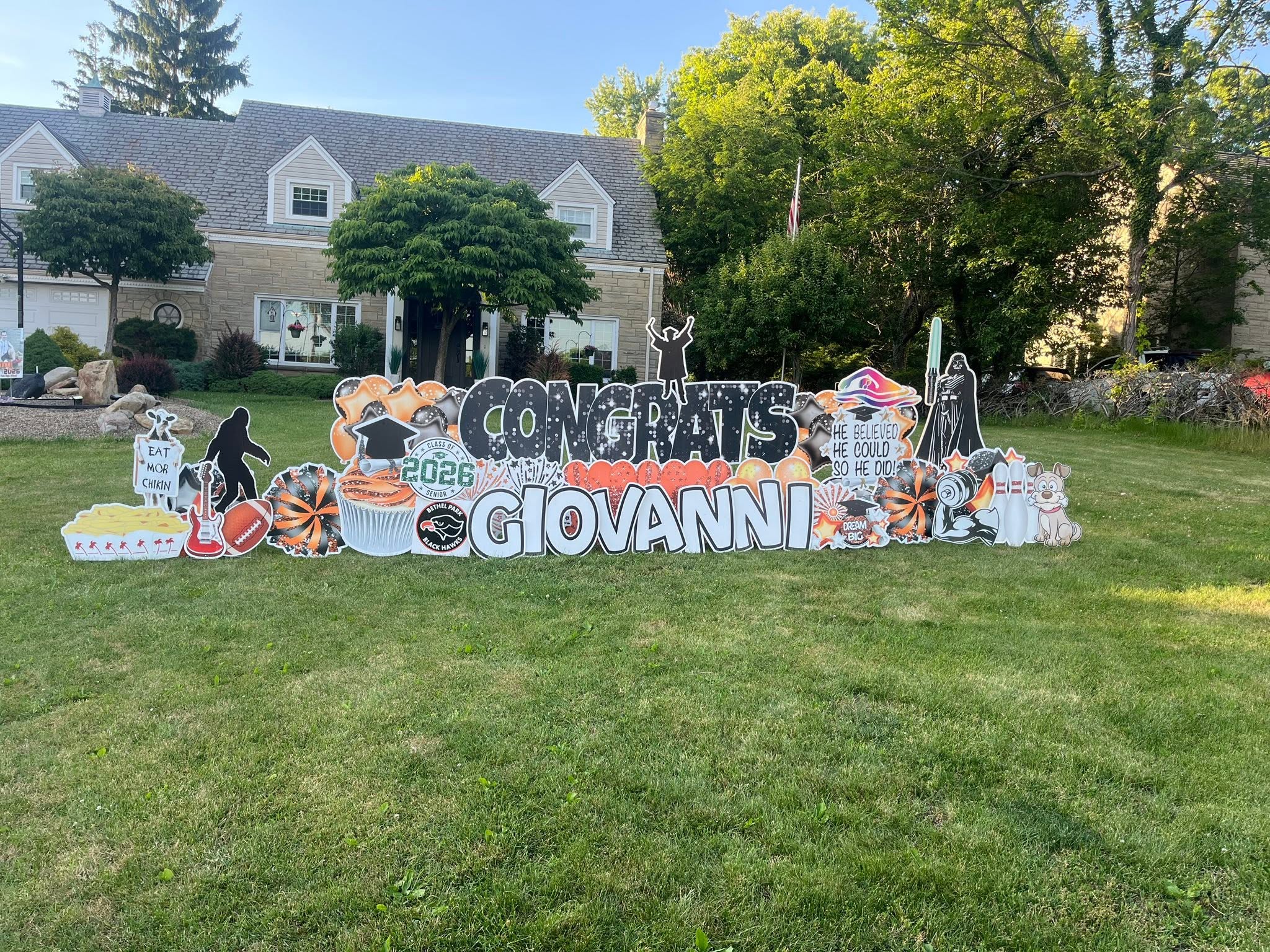 Large outdoor birthday sign with decorations and balloons on a grassy area, houses and trees in the background.