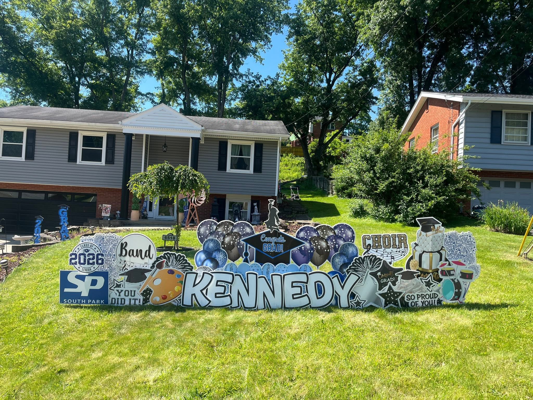 Colorful yard sign with the name 'Kennedy' and various themed decorations in front of houses and trees.