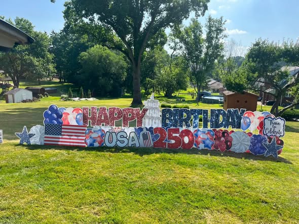 Colorful birthday sign with balloons, stars, and flags on a lawn, celebrating 250th birthday, with trees and houses in background.