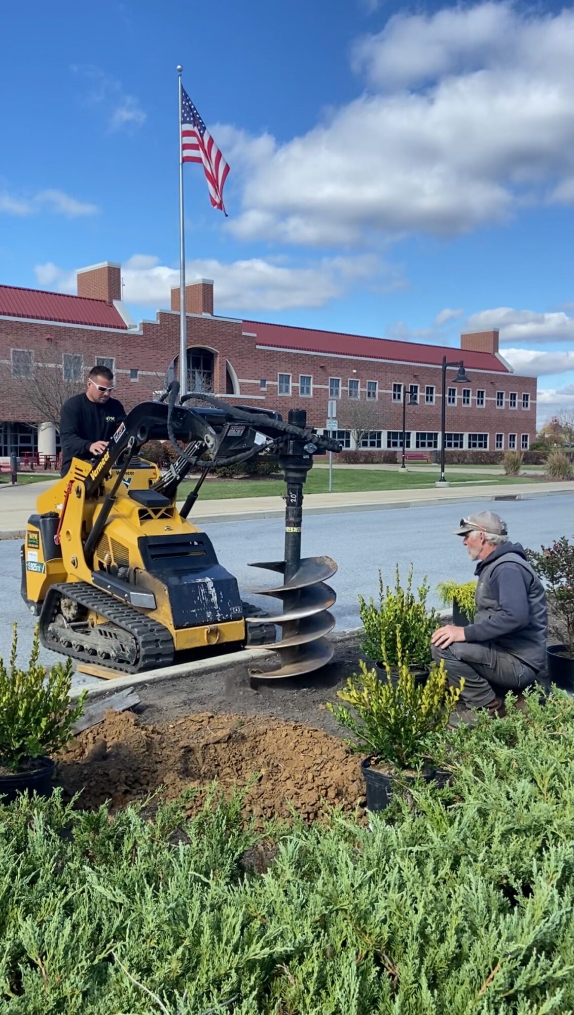 Two people planting a shrub in front of a building with an American flag, blue sky, and clouds, during daytime.