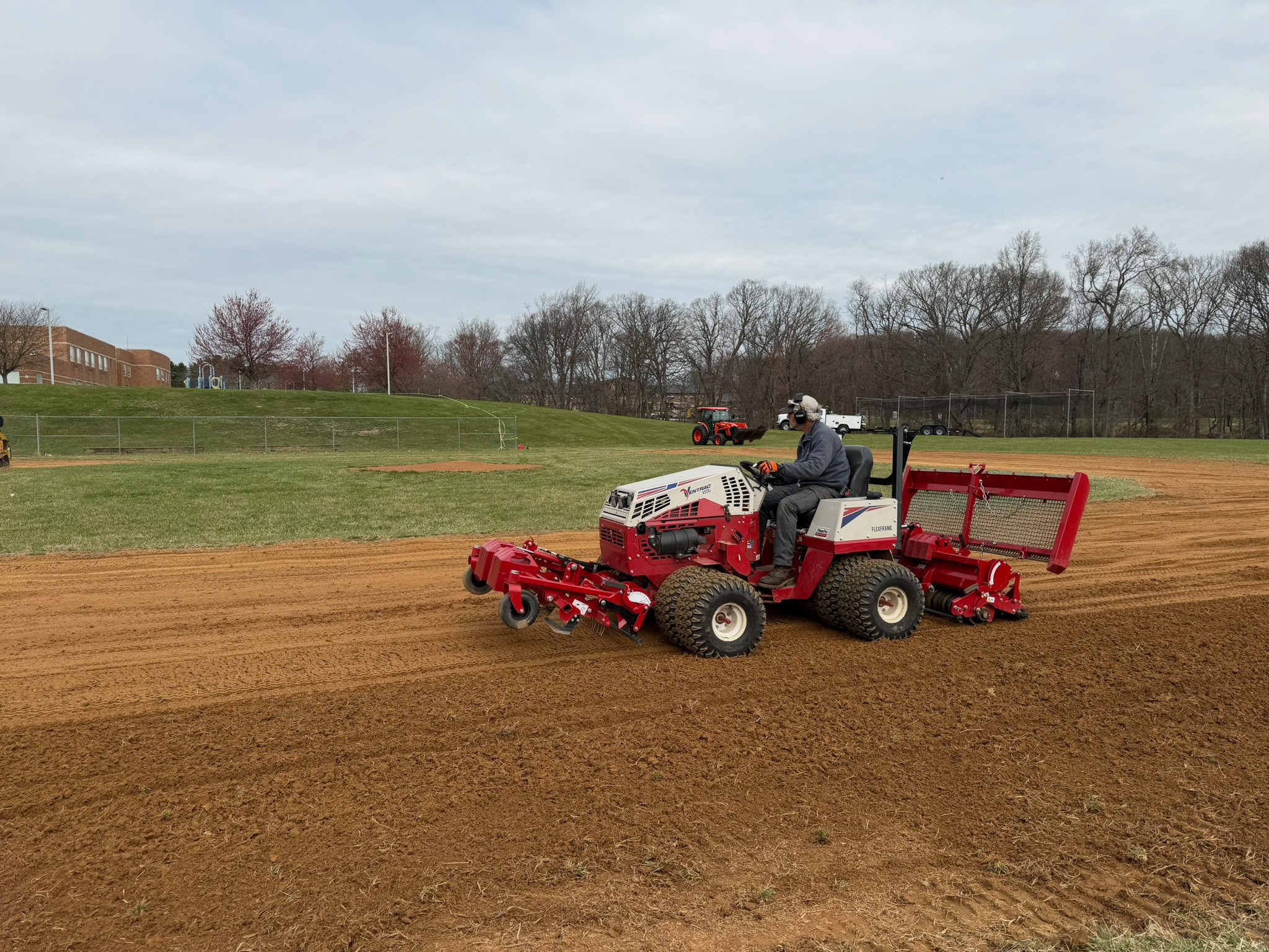 Person operating a red and white riding lawn mower on a dirt field with trees and a building in the background.