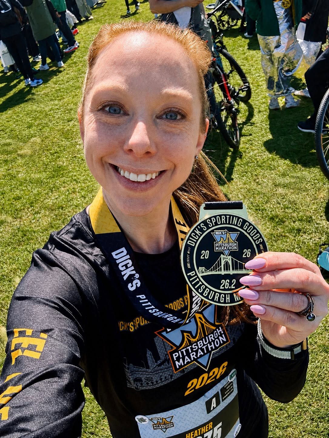 Woman smiling outdoors holding a medal, wearing a black sports jersey with event details, surrounded by people and bicycles on grass.