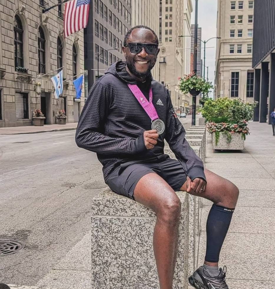 Man sitting on a stone block in an urban street, smiling, wearing sunglasses, a black jacket, shorts, and a medal around his neck.