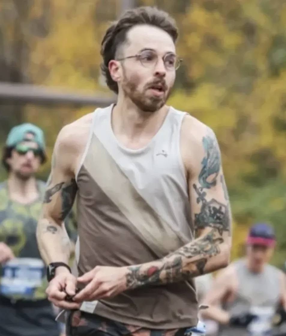 Man with glasses and tattoos running outdoors with trees in background.