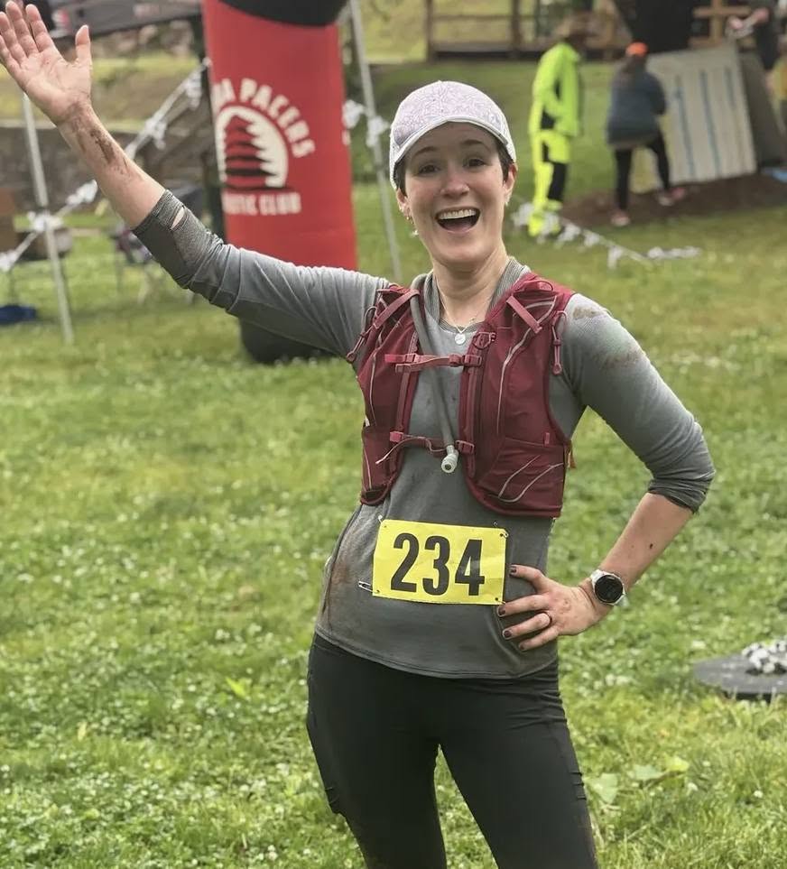 Smiling woman in running gear with race number 234, outdoors on grass, waving, with event banners and people in background.
