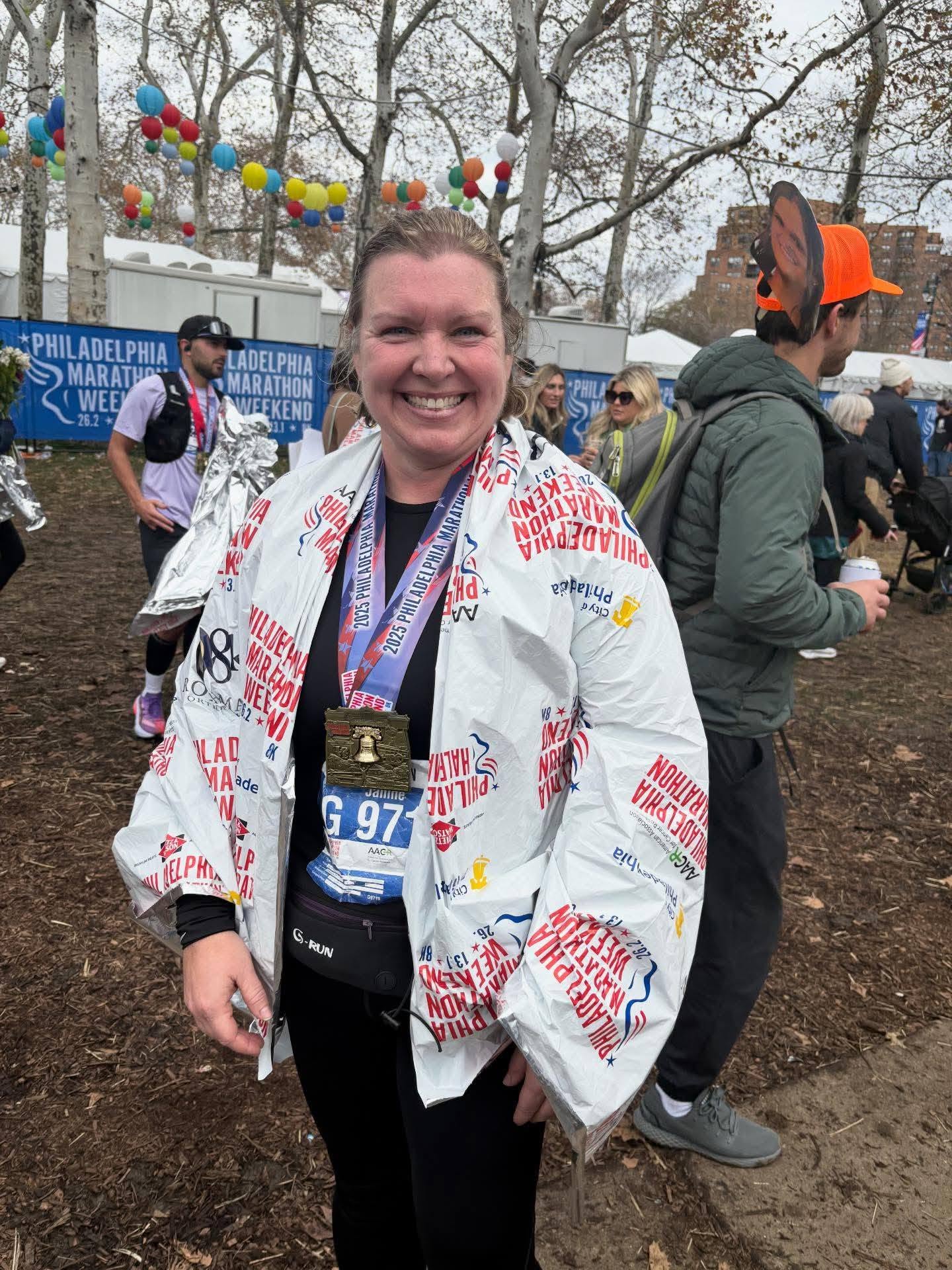Smiling woman wearing a white jacket with red text, medals around her neck, outdoors with trees and people in background.
