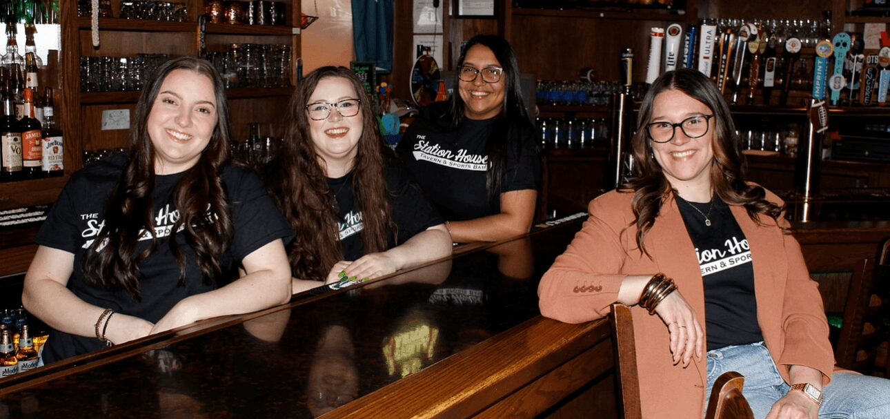 Four women sitting at a bar counter smiling, with bottles and glasses in the background.
