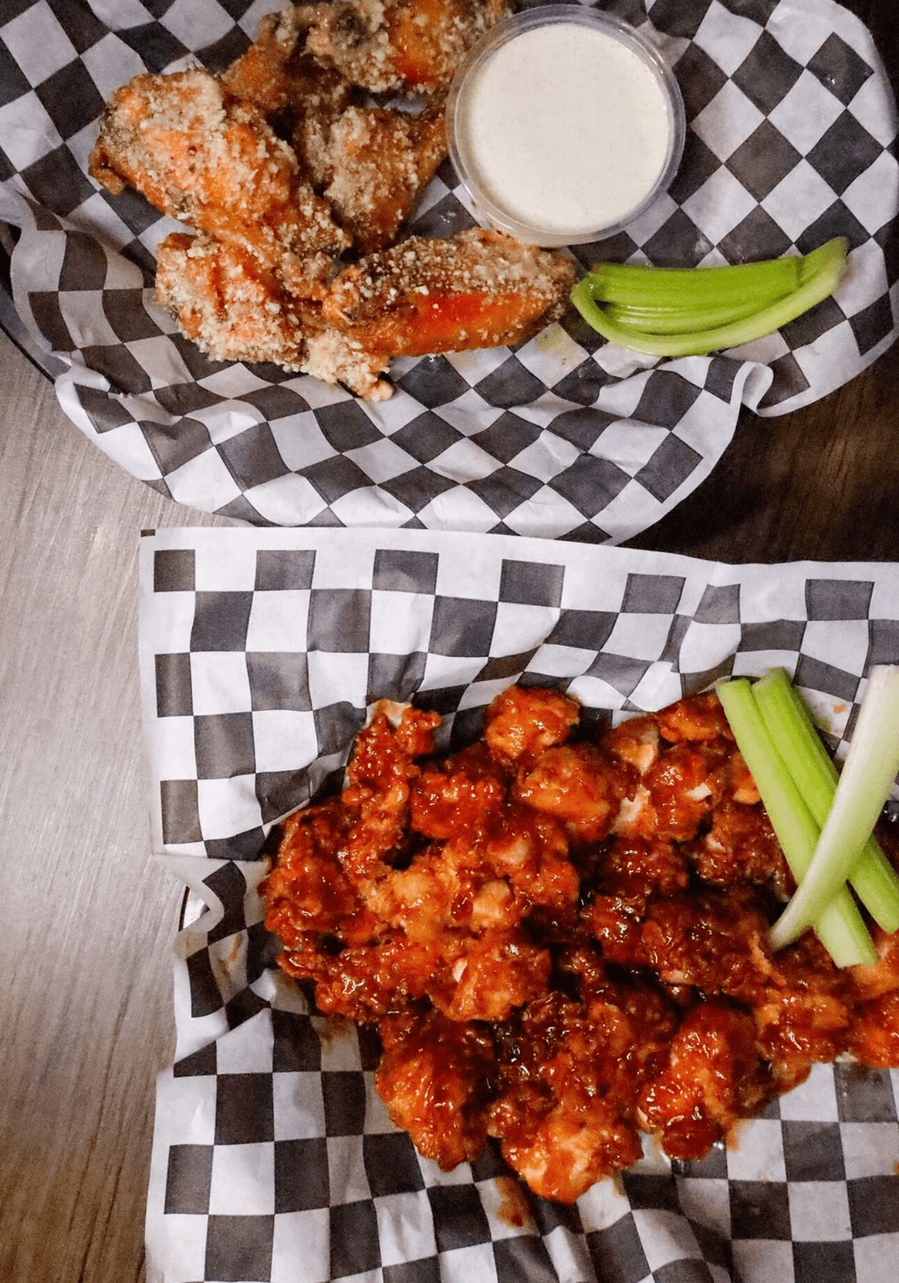 Two baskets of fried chicken with celery sticks and dipping sauces on checkered paper, on a dark wooden table.