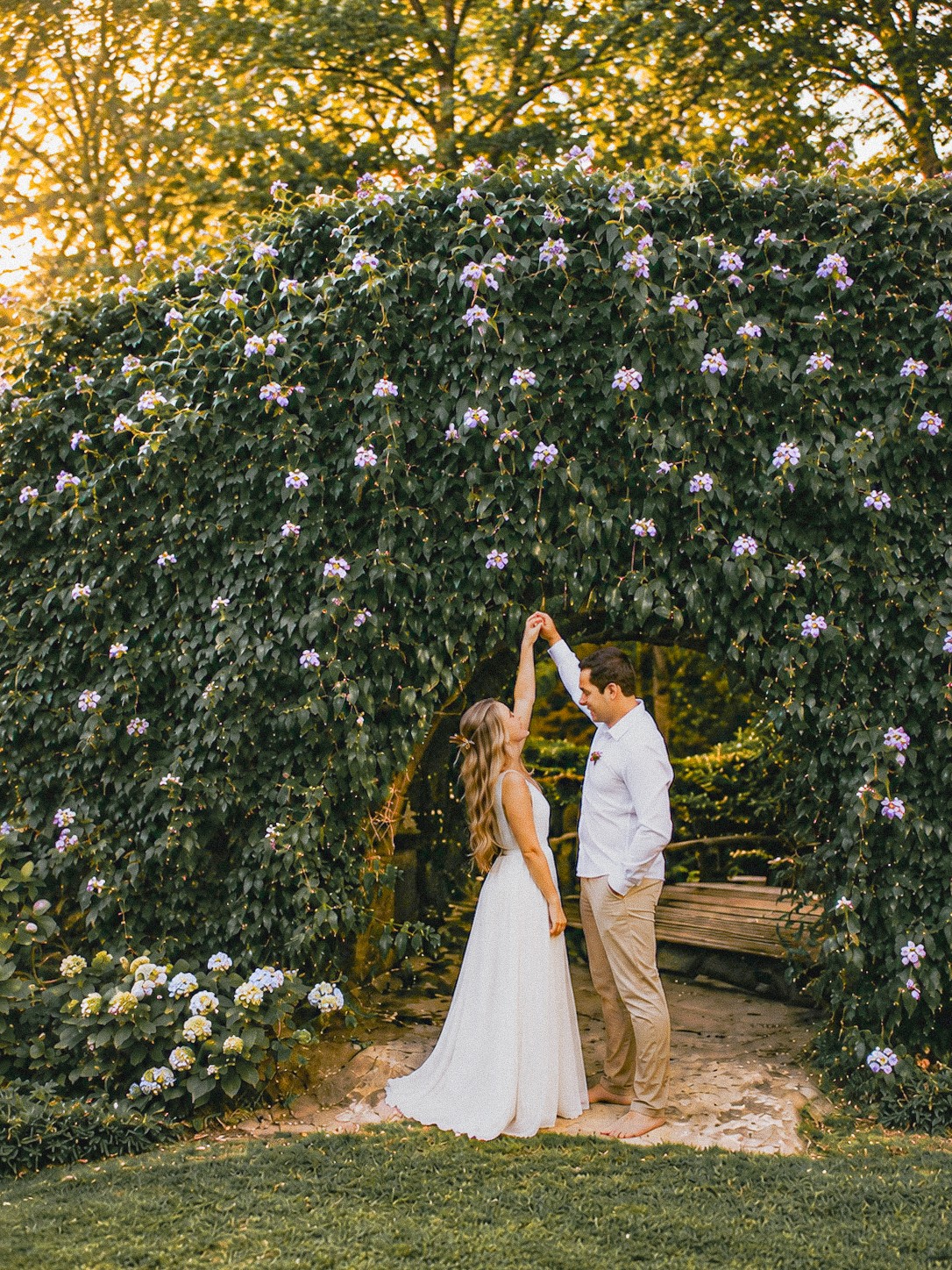 A couple holding hands under a floral arch in a garden during sunset, with trees in the background.