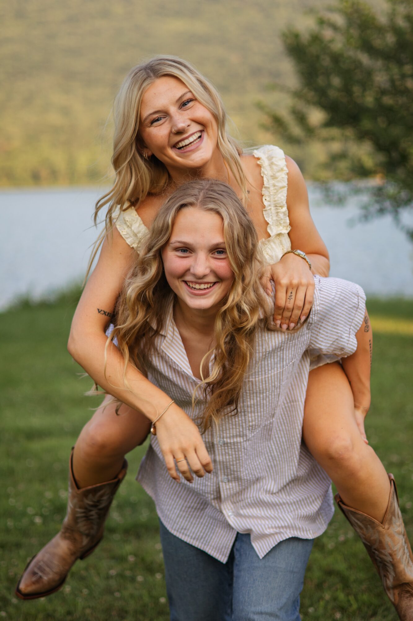 Two smiling women outdoors, one giving the other a piggyback ride, with trees and water in the background.