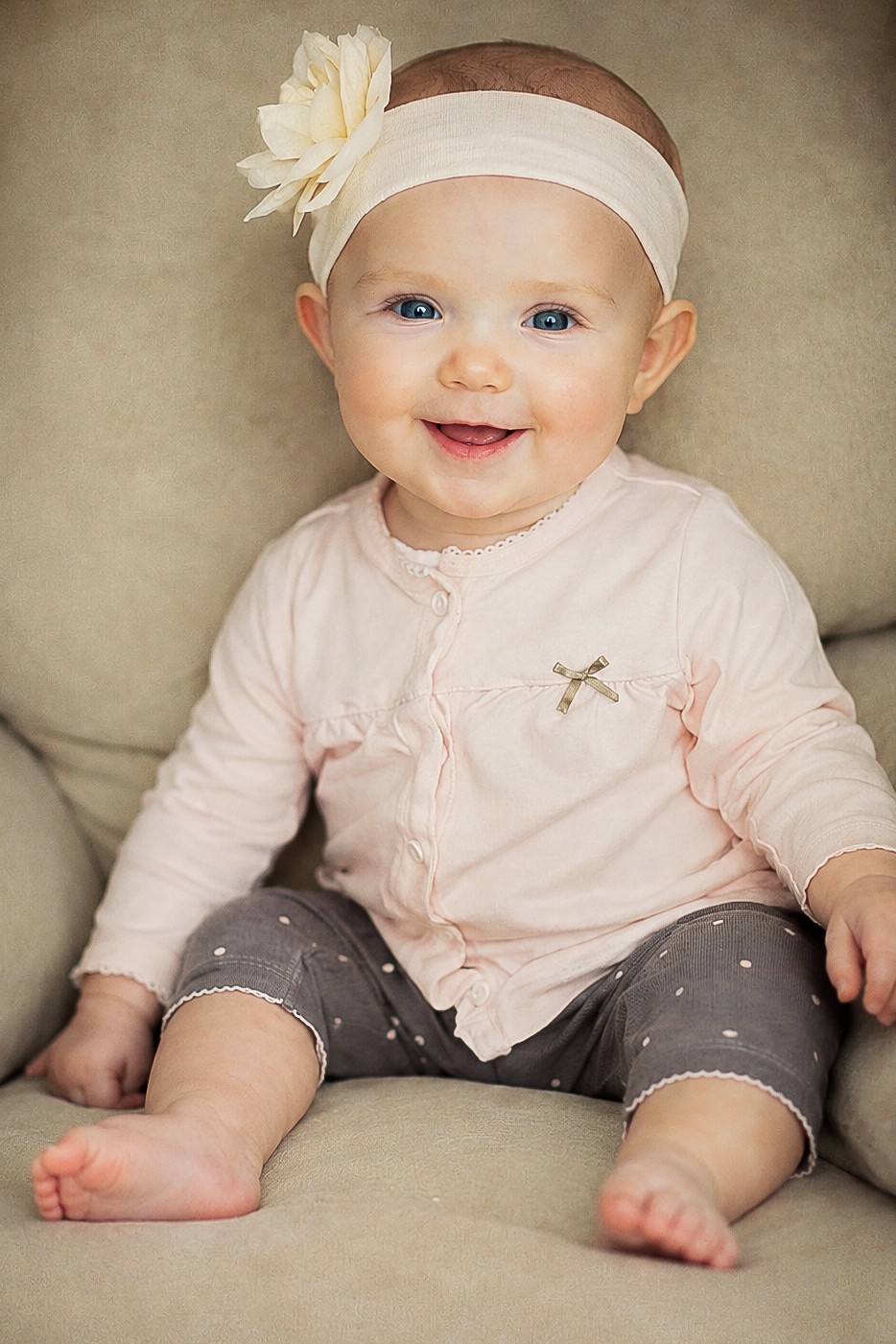 Smiling baby girl with a headband and flower, sitting on a beige couch, wearing a light pink top and gray pants.