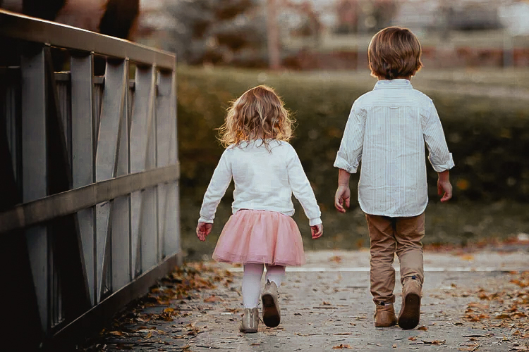 Two children walk away on a path near a fence, with trees and a park in the background.