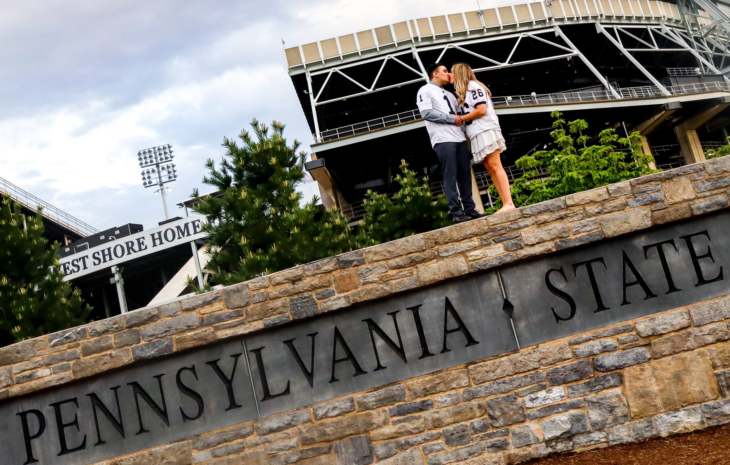 Two people stand on a stone wall with Pennsylvania State University signage, with a stadium and trees in the background.