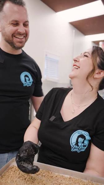 Two healthcare workers smiling and looking at each other in a clinical setting, one woman and one man, both wearing black uniforms with a logo.