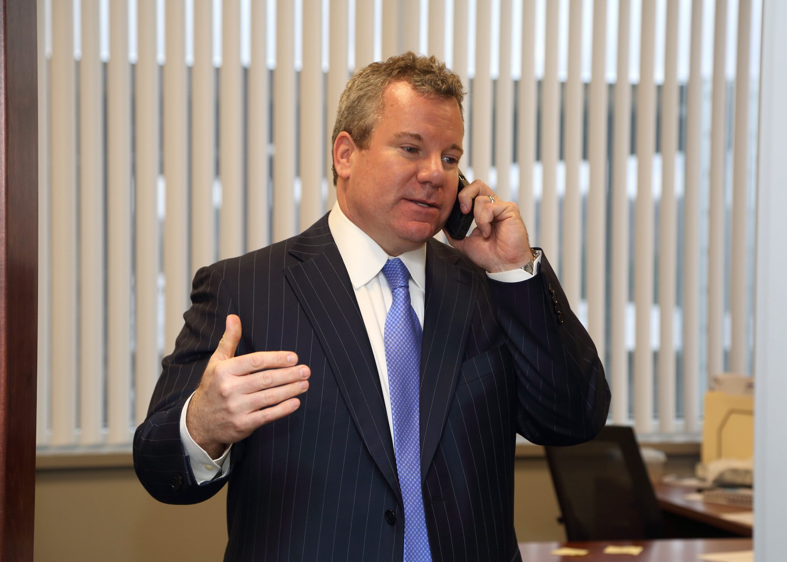 Man in a suit talking on a phone in an office with vertical blinds behind him.
