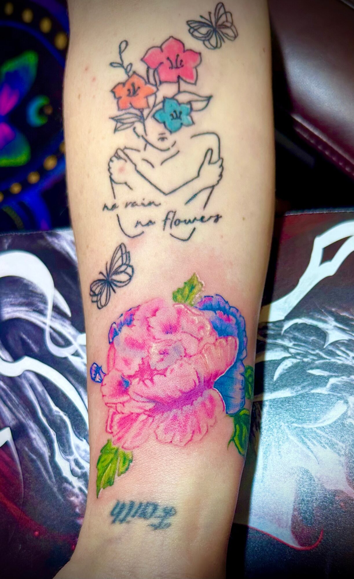 Tattoo of a hand holding flowers, butterflies, and a large pink flower with green leaves, with handwritten text below.