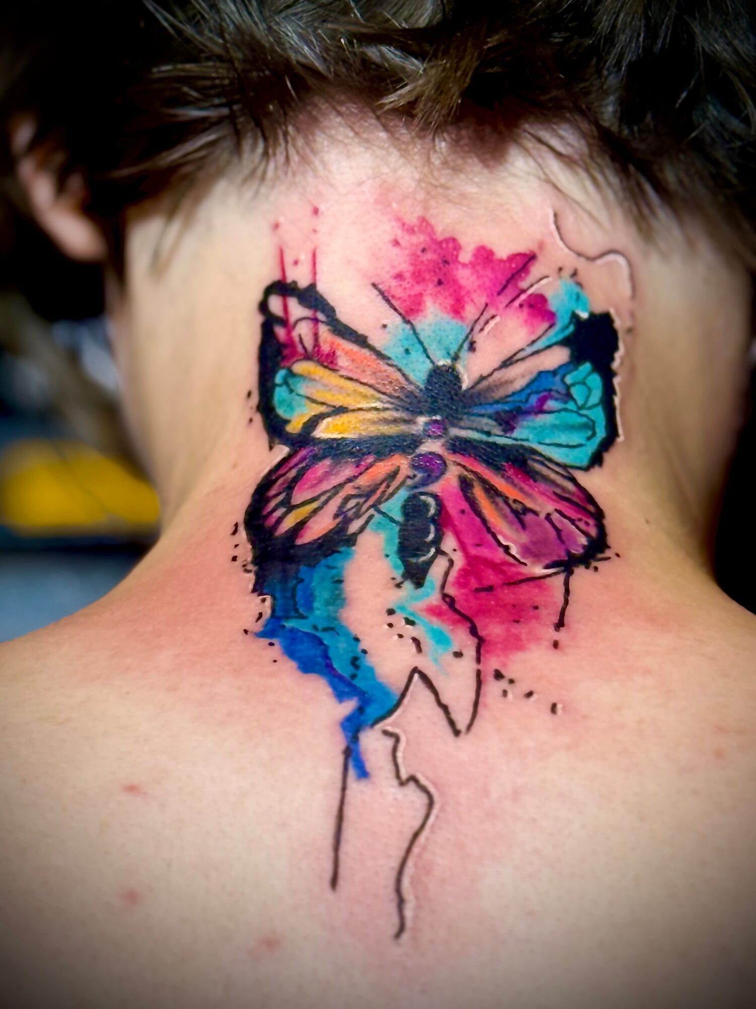 Colorful butterfly tattoo on person's neck with pink, blue, yellow, and purple splashes, and black outlines.
