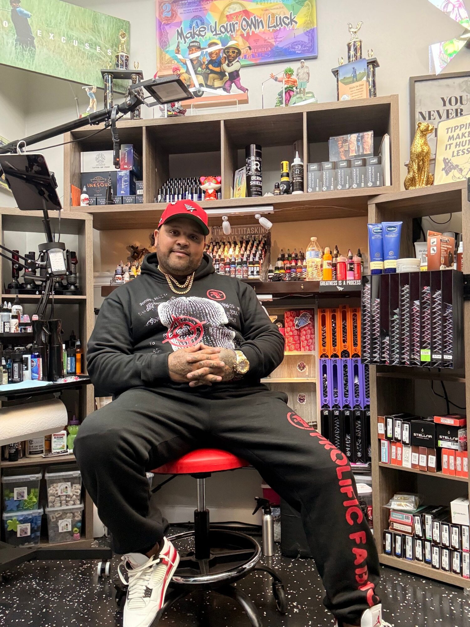 Person sitting on a red stool in a store, surrounded by shelves of products, wearing a black hoodie and black pants.