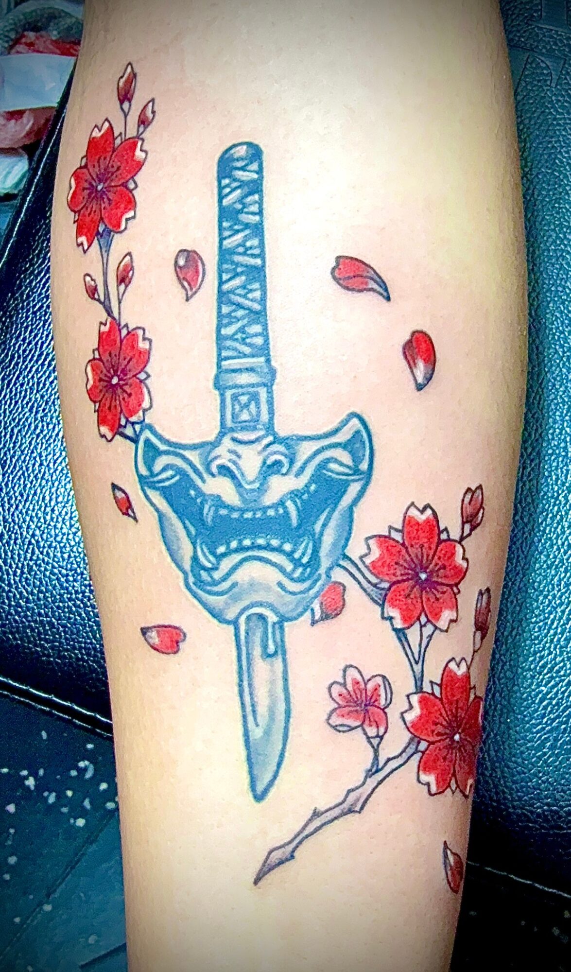 Tattoo of a blue sword with a fierce face, surrounded by red flowers and petals on skin.