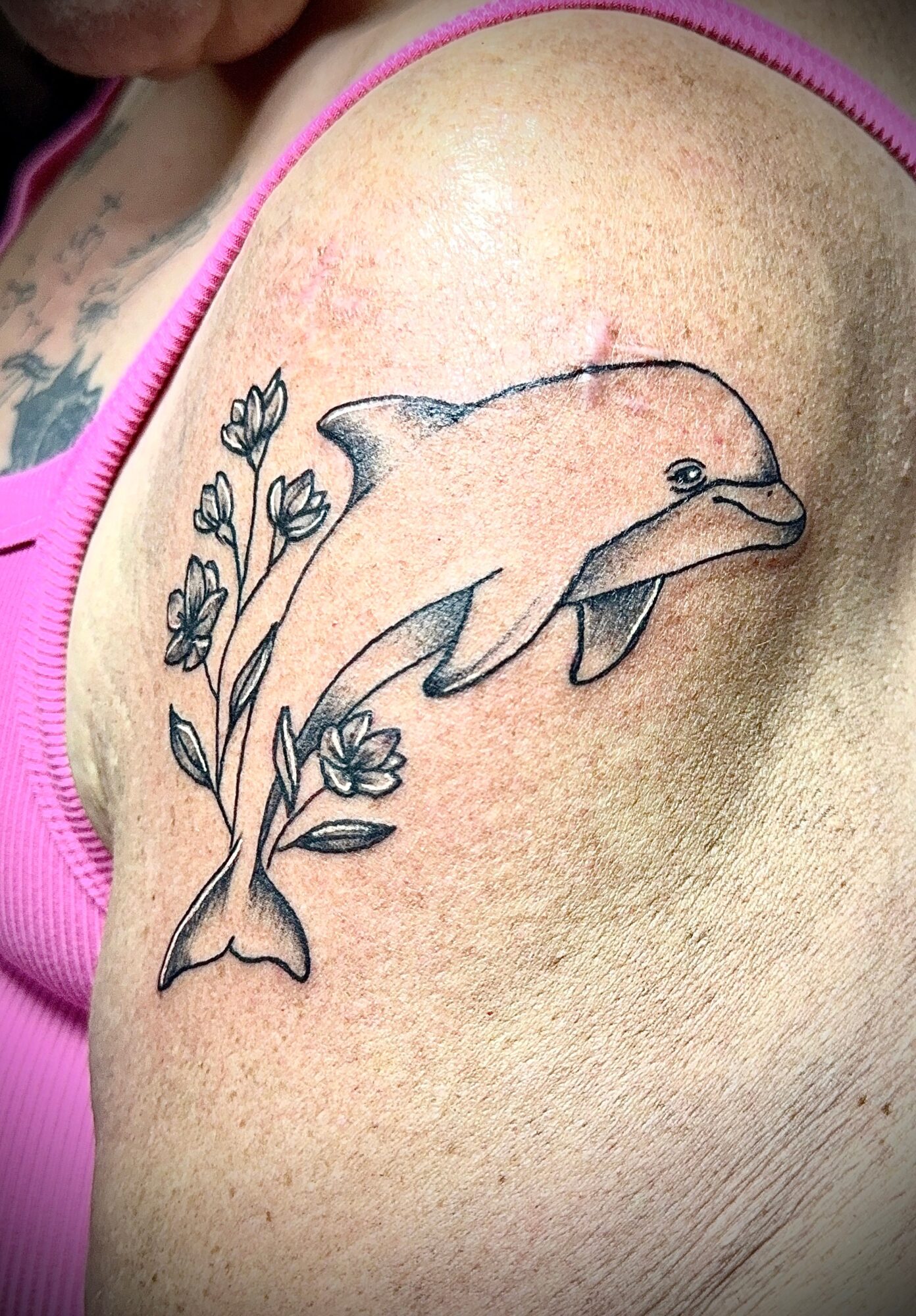 Dolphin jumping over flowers tattoo on shoulder, black outline, detailed design, natural skin background.