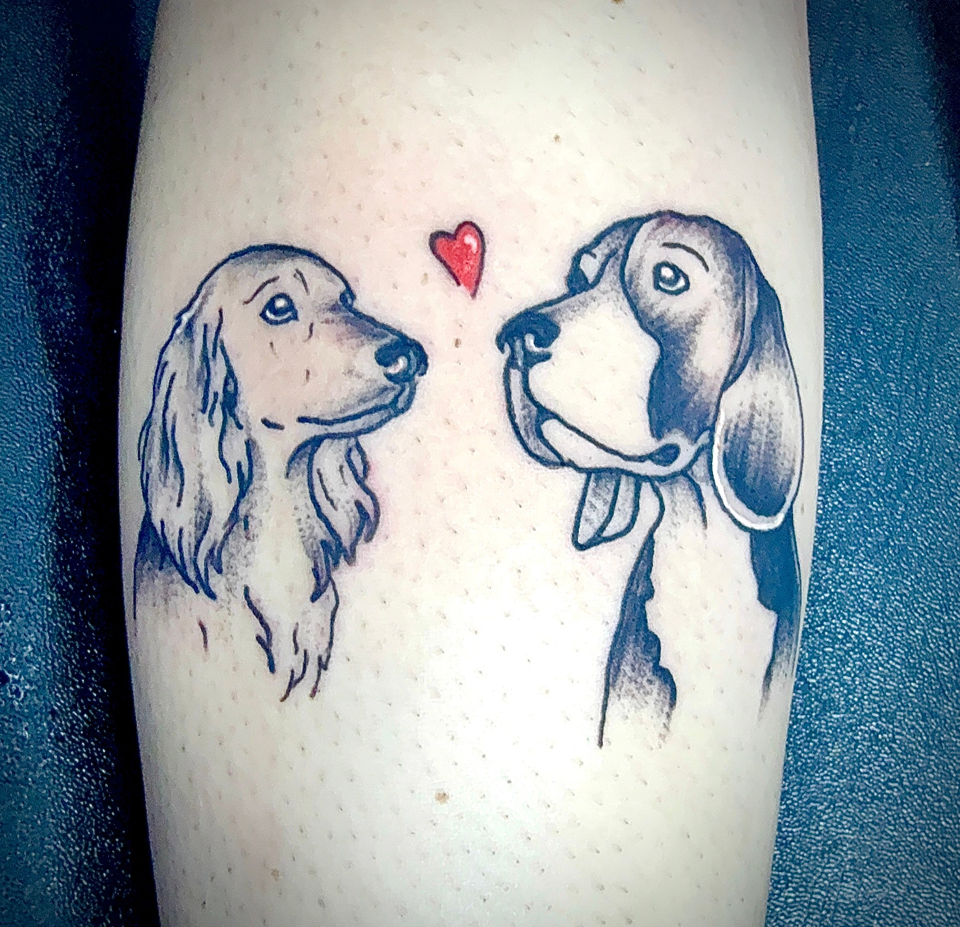 Tattoo of two dogs facing each other with a small red heart above them, in black and gray ink, on skin.