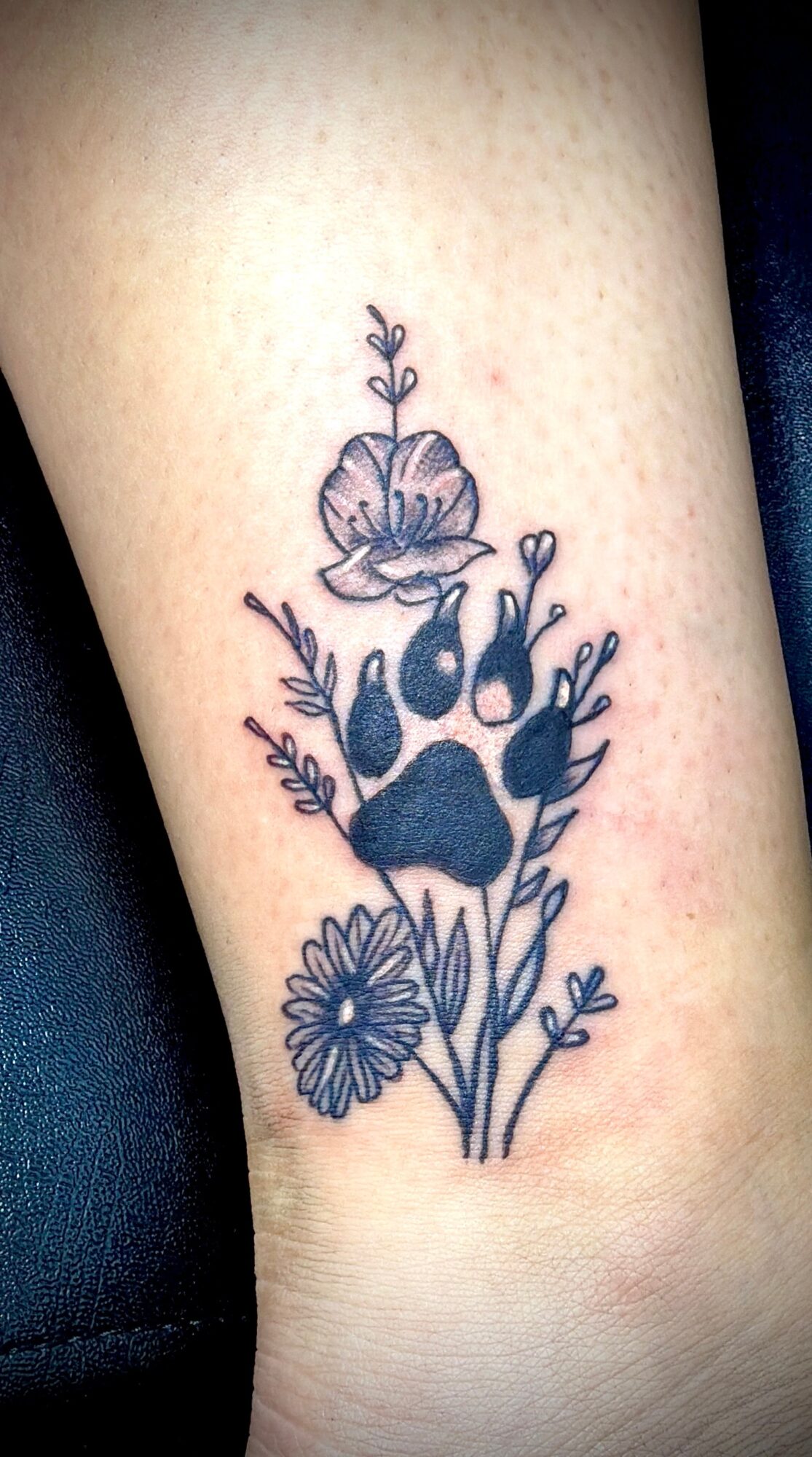 Black floral tattoo with a flower, leaves, and a small daisy on skin.